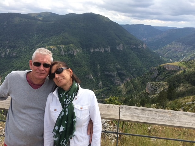 Pascale et Yves Somon, propriétaires souriants du gîte l'Oasis du Lot, posant amoureusement dans leur cadre pittoresque de montagnes verdoyantes.